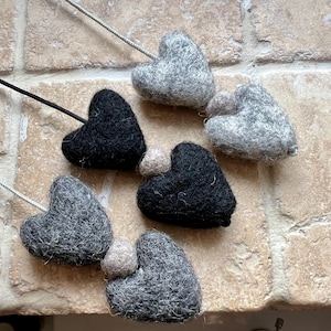 Puede incluir: Adornos de corazón de fieltro hechos a mano en tonos grises y negros. Cada corazón está unido a un cordón fino. Algunos corazones tienen una pequeña bola de color claro adjunta. Los adornos están dispuestos sobre una superficie texturizada.