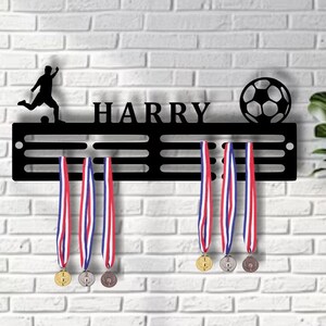 K&ouml;nnte beinhalten: Schwarzer Medaillenhalter aus Metall mit dem Namen "HARRY" und einer Fu&szlig;ball-Silhouette. Der Halter hat 6 F&auml;cher f&uuml;r Medaillen und 3 Medaillen h&auml;ngen daran. Die Medaillen sind in Gold, Silber und Bronze.