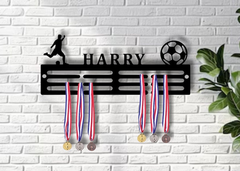 K&ouml;nnte beinhalten: Schwarzer Medaillenhalter aus Metall mit dem Namen "HARRY" und einer Fu&szlig;ball-Silhouette. Der Halter hat 6 F&auml;cher f&uuml;r Medaillen und 3 Medaillen h&auml;ngen daran. Die Medaillen sind in Gold, Silber und Bronze.