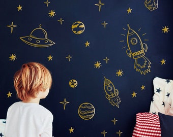 May include: A young child sits on a bed with a white duvet cover and looks at a dark blue wall with gold space-themed decals. The decals include a rocket, a spaceship, planets, stars, and an astronaut. The bed has a red and white checkered pillow and a navy blue pillow.