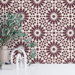 May include: A decorative scene featuring a burgundy and cream geometric patterned wallpaper. A vase with greenery, a white arch, a candle, and a diffuser bottle are on a white surface. The wallpaper has a repeating starburst design.