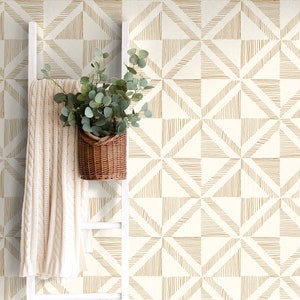 May include: A white wall with a geometric pattern wallpaper featuring a repeating design of brown lines forming squares and triangles. A white ladder leans against the wall with a white blanket draped over it and a potted plant with green leaves in a brown wicker basket.