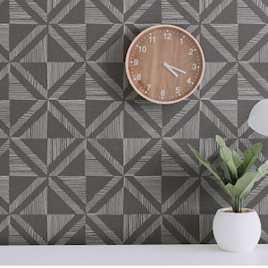 May include: A brown wall with a white geometric pattern. A wooden clock with black hands is hanging on the wall. A white lamp with a white shade is on a table in front of the wall. A potted plant is on the table next to the lamp.