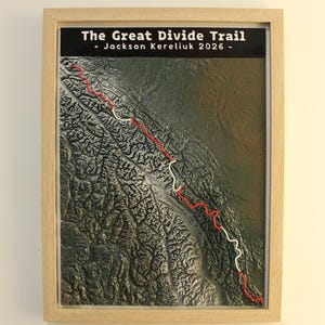 Op de afbeelding: Een ingelijste topografische kaart getiteld The Great Divide Trail - Jackson Kereliuk 2026. De kaart toont een donker, getextureerd berglandschap met een opvallend pad gemarkeerd door afwisselende rode en witte lijnen over het oppervlak.
