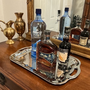 May include: A rectangular silver-toned serving tray with scalloped edges and two handles. The tray holds a clear bottle of Pinnacle Vodka, a square bottle of Flor de Caña 12 rum, and a dark bottle of blackberry liqueur, all arranged on a wooden surface.
