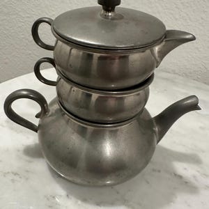 May include: A stackable, antique pewter tea set. The set includes a teapot with a spout and handle, two cups, and a lid with a small, round knob. The pieces are arranged on top of each other, creating a compact design.