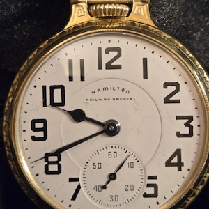 May include: A gold-tone Hamilton Railway Special pocket watch with a white face and black numerals. The watch has a subdial for seconds and black hands. The watch is in a gold-tone case with a decorative design.
