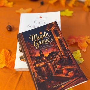 Può includere: Un libro intitolato Maple Grove di Misa Slowliving con un'illustrazione di copertina scura di una città al tramonto, con zucche su un portico e foglie autunnali. Dietro si trova un libro bianco intitolato Caffe, Sogni, con una foglia d'acero.
