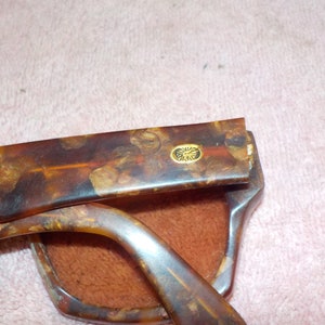 Puede incluir: Un par de gafas de sol de carey con un logotipo dorado en el lateral. Las gafas est&aacute;n plegadas y se ve la funda.