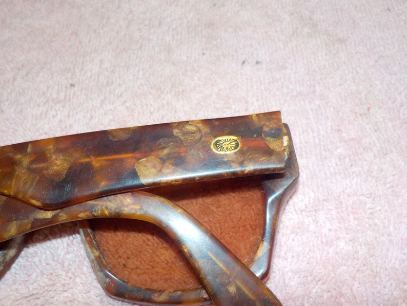 Puede incluir: Un par de gafas de sol de carey con un logotipo dorado en el lateral. Las gafas est&aacute;n plegadas y se ve la funda.