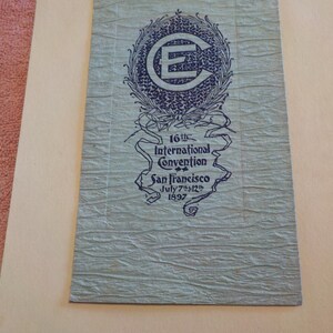 Puede incluir: Papel azul verdoso vintage con el texto "16th International Convention San Francisco July 7th-12th 1897". El papel tiene una superficie texturizada y un emblema azul oscuro con la letra "E".