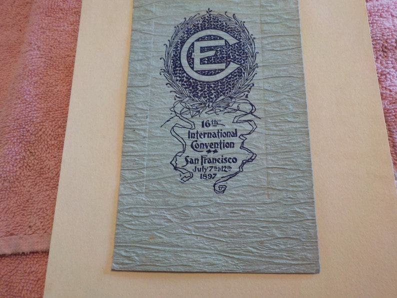 Puede incluir: Papel azul verdoso vintage con el texto "16th International Convention San Francisco July 7th-12th 1897". El papel tiene una superficie texturizada y un emblema azul oscuro con la letra "E".