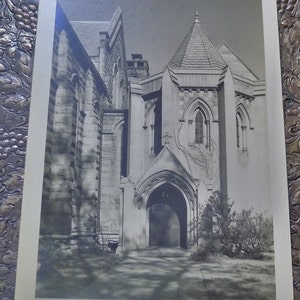 May include: A black and white photograph of a stone church building with a pointed archway entrance. The building has a tall, pointed tower and intricate stonework.