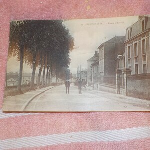 May include: A black and white vintage postcard showing a street scene in Neufchateau, France. The street is lined with trees and there are buildings on both sides. Two people are walking down the street.