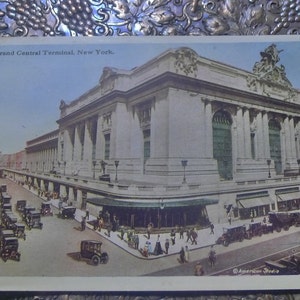 May include: A vintage postcard depicting Grand Central Terminal in New York City. The postcard shows the exterior of the building, with its iconic clock tower and arched windows. The street in front of the terminal is filled with cars and people.