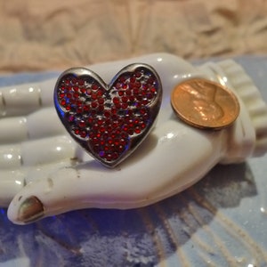 Puede incluir: Un anillo de plata con un diseño de corazón de diamantes de imitación rojos.