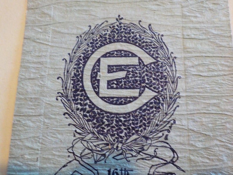 Puede incluir: Papel azul claro texturizado con un emblema azul oscuro. El emblema presenta una gran letra "E" dentro de un c&iacute;rculo con una flecha, rodeada por una corona floral. El texto "16th" es visible en la parte inferior.