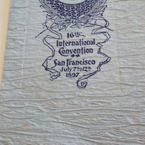 Puede incluir: Papel texturizado azul claro con texto azul oscuro. El texto dice "16&ordf; Convenci&oacute;n Internacional San Francisco 7 al 12 de julio de 1897". La parte superior del papel tiene un dise&ntilde;o floral decorativo.