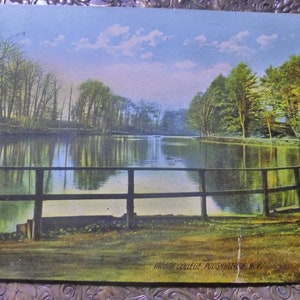 May include: A vintage postcard depicting a scenic lake with a wooden fence in the foreground. The lake is surrounded by lush green trees and a blue sky with white clouds. The text "Wesley College, Rochester, N.Y." is printed at the bottom of the postcard.