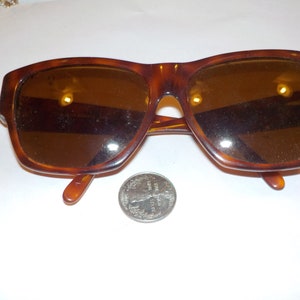 May include: Brown tortoiseshell sunglasses with rectangular frames. The sunglasses have dark lenses and a classic design. A silver coin is next to the sunglasses.