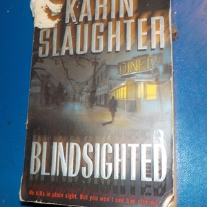 Könnte beinhalten: Buchcover für den Roman "Blindsighted" von Karin Slaughter. Das Cover zeigt eine dunkle Nachtszene einer Straße mit einem Diner im Hintergrund. Der Titel "Blindsighted" ist in großen weißen Buchstaben geschrieben.