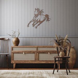 Op de afbeelding: Een bronzen metalen wandkunstwerk dat een rennend paard afbeeldt. Het paard staat in volle galop, met uitgestrekte benen en manen in de wind. Het kunstwerk is gemonteerd op een grijze wand met panelen, boven een houten kast met geweven panelen.