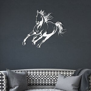 Op de afbeelding: Een witte metalen wandkunst die een rennend paard afbeeldt. Het paard is in volle galop, met uitgestrekte benen en wapperende manen. Het kunstwerk is bevestigd aan een donkergrijze muur, boven een bank met patroon.