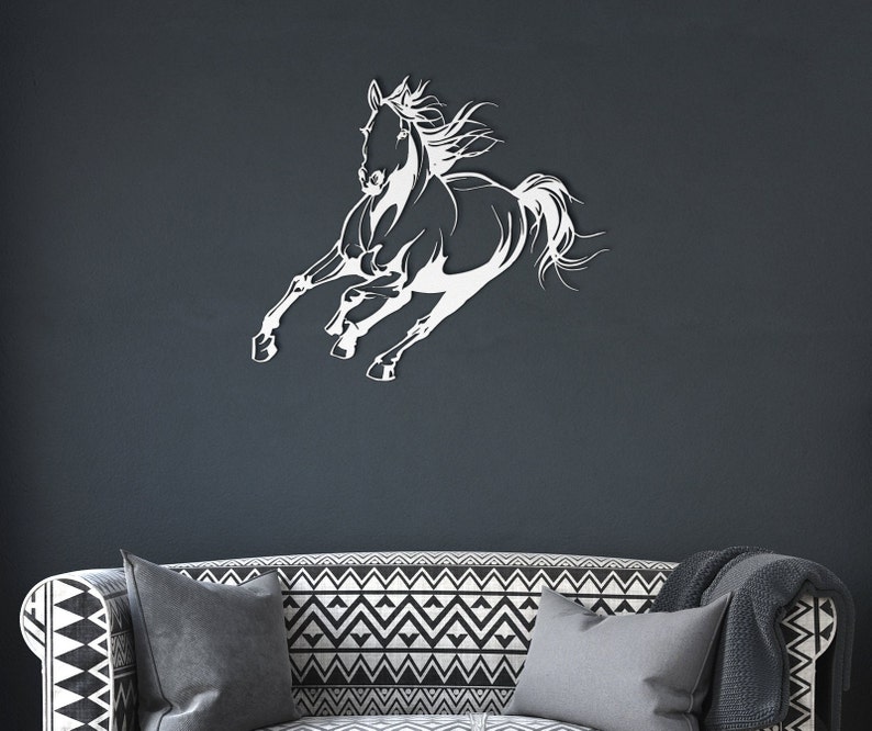 Op de afbeelding: Een witte metalen wandkunst die een rennend paard afbeeldt. Het paard is in volle galop, met uitgestrekte benen en wapperende manen. Het kunstwerk is bevestigd aan een donkergrijze muur, boven een bank met patroon.