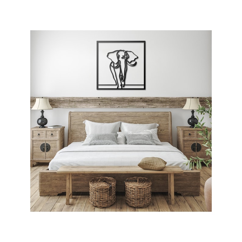 K&ouml;nnte beinhalten: Eine schwarze Wandskulptur aus Metall, die einen Elefanten in einem einfachen Line-Art-Stil darstellt. Die Skulptur h&auml;ngt &uuml;ber einem Holzbettgestell mit wei&szlig;en Bettw&auml;sche und einer Holzbank.