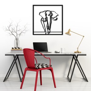 K&ouml;nnte beinhalten: Eine schwarze Wandskulptur aus Metall, die einen Elefanten im minimalistischen Line-Art-Stil darstellt. Die Skulptur h&auml;ngt an einer wei&szlig;en Wand &uuml;ber einem Schreibtisch mit einem roten Stuhl, einem Laptop und einer Lampe.