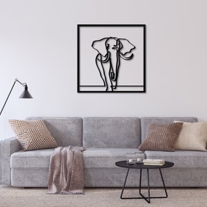 K&ouml;nnte beinhalten: Eine Schwarz-Wei&szlig;-Linienzeichnung eines Elefanten, gerahmt in einem schwarzen quadratischen Rahmen, h&auml;ngt an einer wei&szlig;en Wand &uuml;ber einem grauen Sofa mit Kissen und einer Decke.