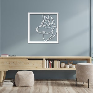 Pode incluir: Uma moldura de madeira branca com um desenho de linha da cabe&ccedil;a de um c&atilde;o de perfil. O c&atilde;o est&aacute; olhando para a direita e tem um focinho longo e orelhas ca&iacute;das. A moldura est&aacute; pendurada em uma parede azul claro acima de uma prateleira de madeira com alguns livros e uma planta em vaso.