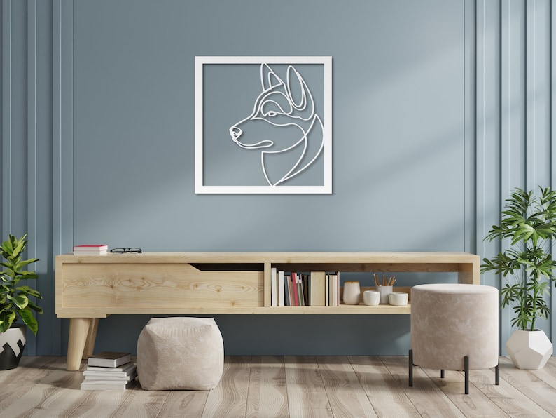 Pode incluir: Uma moldura de madeira branca com um desenho de linha da cabe&ccedil;a de um c&atilde;o de perfil. O c&atilde;o est&aacute; olhando para a direita e tem um focinho longo e orelhas ca&iacute;das. A moldura est&aacute; pendurada em uma parede azul claro acima de uma prateleira de madeira com alguns livros e uma planta em vaso.