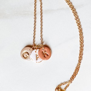 May include: A gold chain necklace with three round ceramic charms. The charms are in shades of beige, white, and pink, and each has a gold letter on it: G, A, and K.