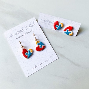 May include: A pair of gold heart-shaped earrings with a blue, red, and yellow puzzle piece design. The earrings are displayed on a white card with the text "A Little Selah Handcrafted Designs" and "made just for you".