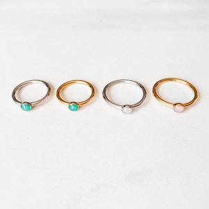 May include: Four delicate rings with small, round gemstones. Two rings are silver with a turquoise and a white gemstone. The other two rings are gold, one with a turquoise stone and the other with a pink stone. All rings are on a white surface.