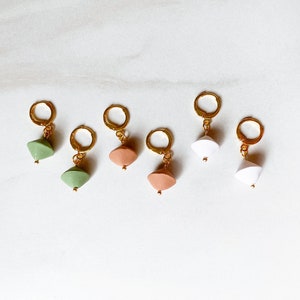 May include: Five pairs of gold hoop earrings with different colored geometric charms. The charms are green, peach, and white.