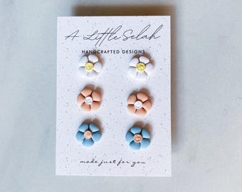Puede incluir: Un conjunto de tres pares de pendientes de flores de arcilla polimérica hechos a mano. Los pendientes son de color blanco, melocotón y azul con centros amarillos. La tarjeta dice "A Little Selah Handcrafted Designs made just for you."