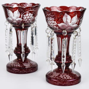 May include: Two ruby red glass candle holders with etched grape and leaf designs. Each holder has a scalloped rim and dangling crystal prisms. The bases and bowls are decorated with etched grape and leaf patterns.