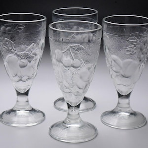 May include: Four clear, stemmed glasses with etched fruit designs. Each glass features a wide, circular base, a slender stem, and a flared bowl. The etched designs include apples, cherries, and other fruits. The glasses are ideal for serving beverages.