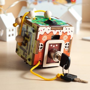 May include: A wooden activity cube featuring a gingerbread house design with a brown, white, and red color scheme. One side has a metal key lock with a silver key attached by a yellow cord. Another side shows a green maze with wooden beads on a cord.