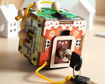 May include: A wooden activity cube featuring a gingerbread house design with a brown, white, and red color scheme. One side has a metal key lock with a silver key attached by a yellow cord. Another side shows a green maze with wooden beads on a cord.