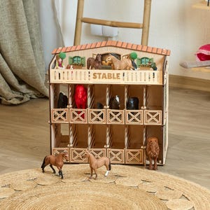 May include: A two-storey wooden toy stable with a tiled roof and the word STABLE printed on the front. The structure features multiple stalls containing various plastic horse figurines in black, brown, and red, with additional horses standing in front.