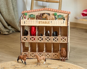 Op de afbeelding: Een houten speelgoedstal met twee verdiepingen, een pannendak en het woord STABLE op de voorkant gedrukt. De structuur heeft meerdere stallen met diverse plastic paardenfiguren in zwart, bruin en rood, met extra paarden die ervoor staan.
