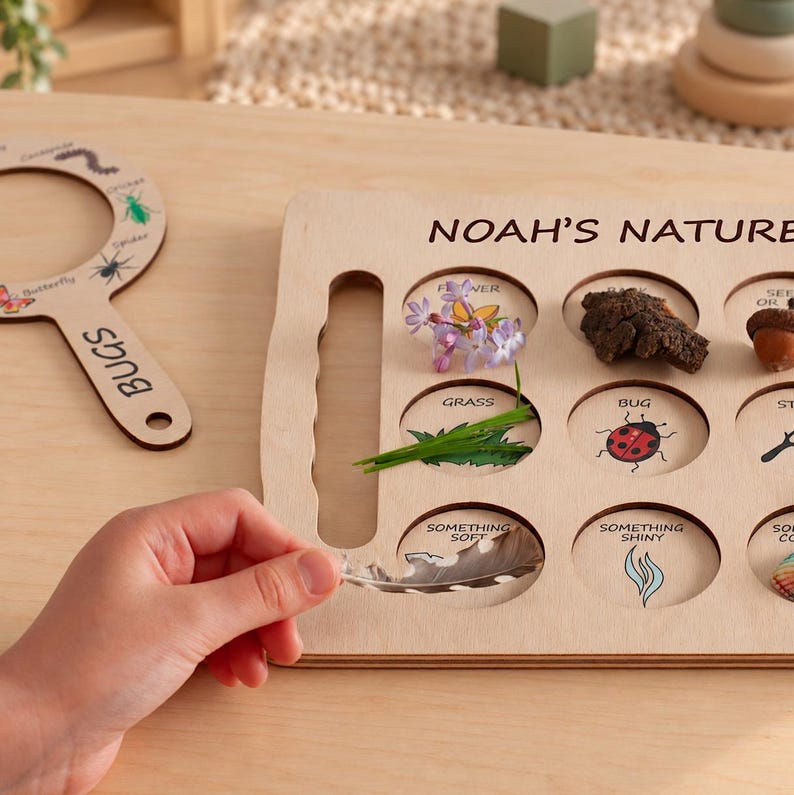 May include: A light-colored wooden nature scavenger hunt board labeled NOAH'S NATURE with circular cutouts containing natural items like flowers, bark, grass, and a feather, alongside a matching wooden magnifying glass labeled BUGS.
