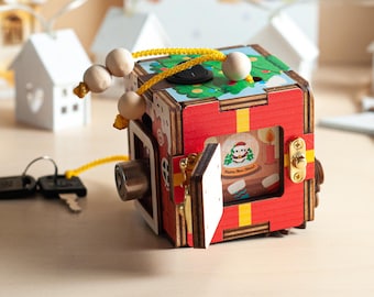 May include: A wooden cube-shaped activity box with a red exterior, yellow stripes, and a hinged door revealing a graphic of a panda in a snow globe with the text Happy New Year. It features a metal lock, keys on a yellow cord, and wooden beads.