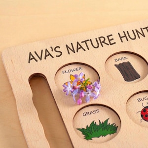 Wooden educational toy with the words "NATURE HUB" printed on it. The toy has cutouts labeled "FLOWER", "BARK", "GRASS", and "BUG". A small bunch of purple flowers is placed in the "FLOWER" cutout.