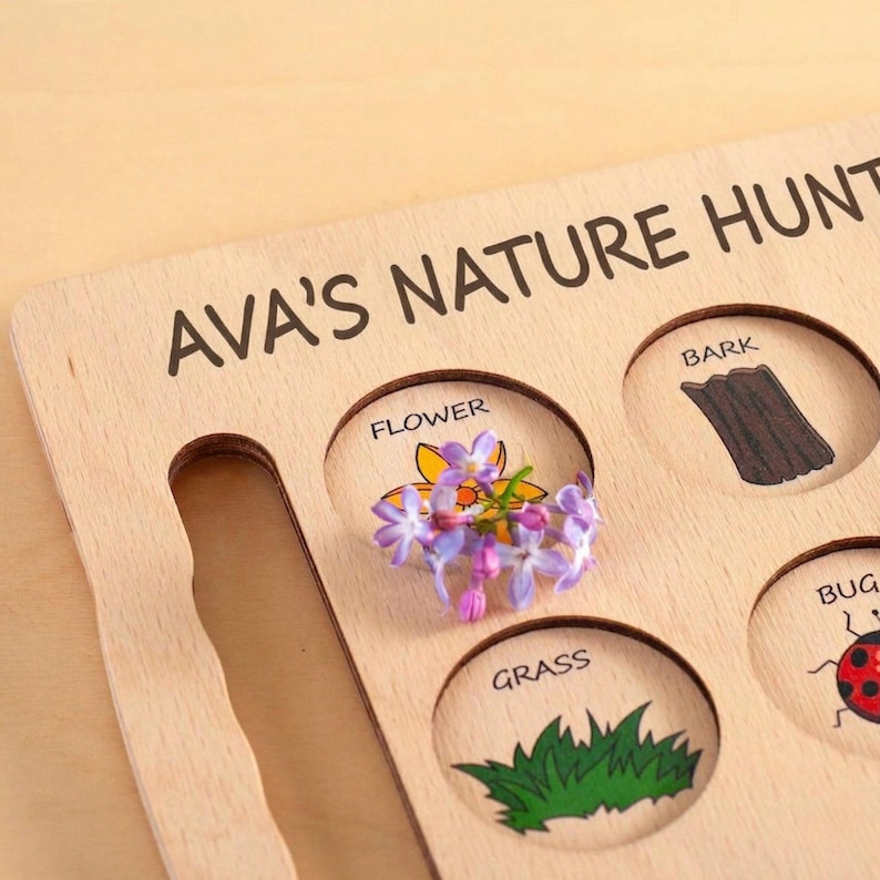 Wooden educational toy with the words "NATURE HUB" printed on it. The toy has cutouts labeled "FLOWER", "BARK", "GRASS", and "BUG". A small bunch of purple flowers is placed in the "FLOWER" cutout.