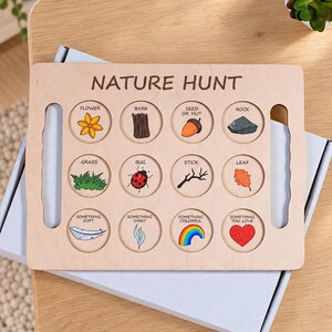 May include: A rectangular light-colored wooden nature hunt board featuring the text NATURE HUNT at the top. It contains twelve circular cutouts with illustrations and labels for items like a flower, bark, seed, rock, grass, bug, stick, leaf, and more.