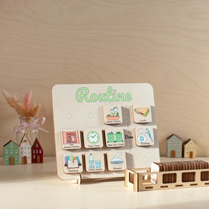 Op de afbeelding: Houten routinebord met groene tekst "Routine" en ge&iuml;llustreerde taaktegels. Taken zijn onder meer "Water Plants", "Lunch", "Put Away Clothes" en "Flossing". Een houten opbergdoos bevat de taaktegels. Miniatuurhuisjes op de achtergrond.